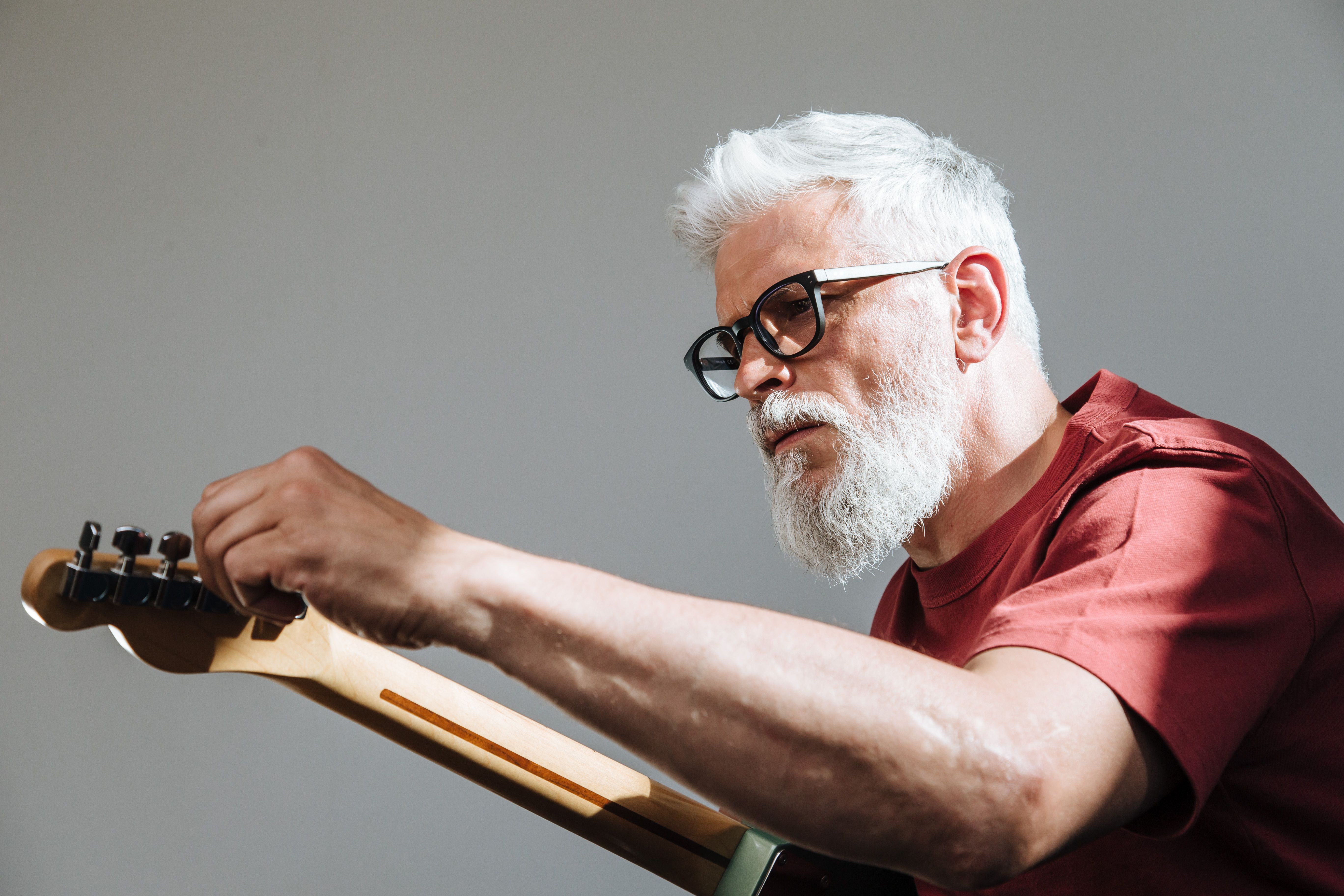 Older man with white beard and glasses carefully tuning an acoustic guitar — focused and deliberate at any age
